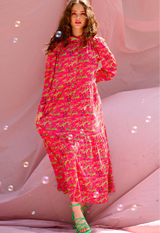 Woman standing in a pink dress and green shoes looking into the camera, the background is a pink flowy sheet.