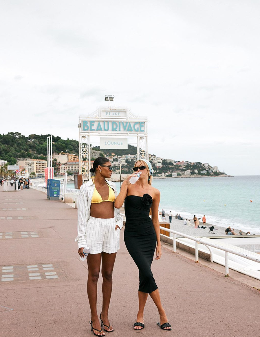 Two women standing on a boulevard, on the right side of the image you can see the ocean.