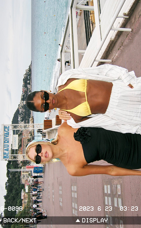 Two woman standing on a boulevard. The one on the left is wearing a black dress and the woman to the roight is wearing a yellow bikini.